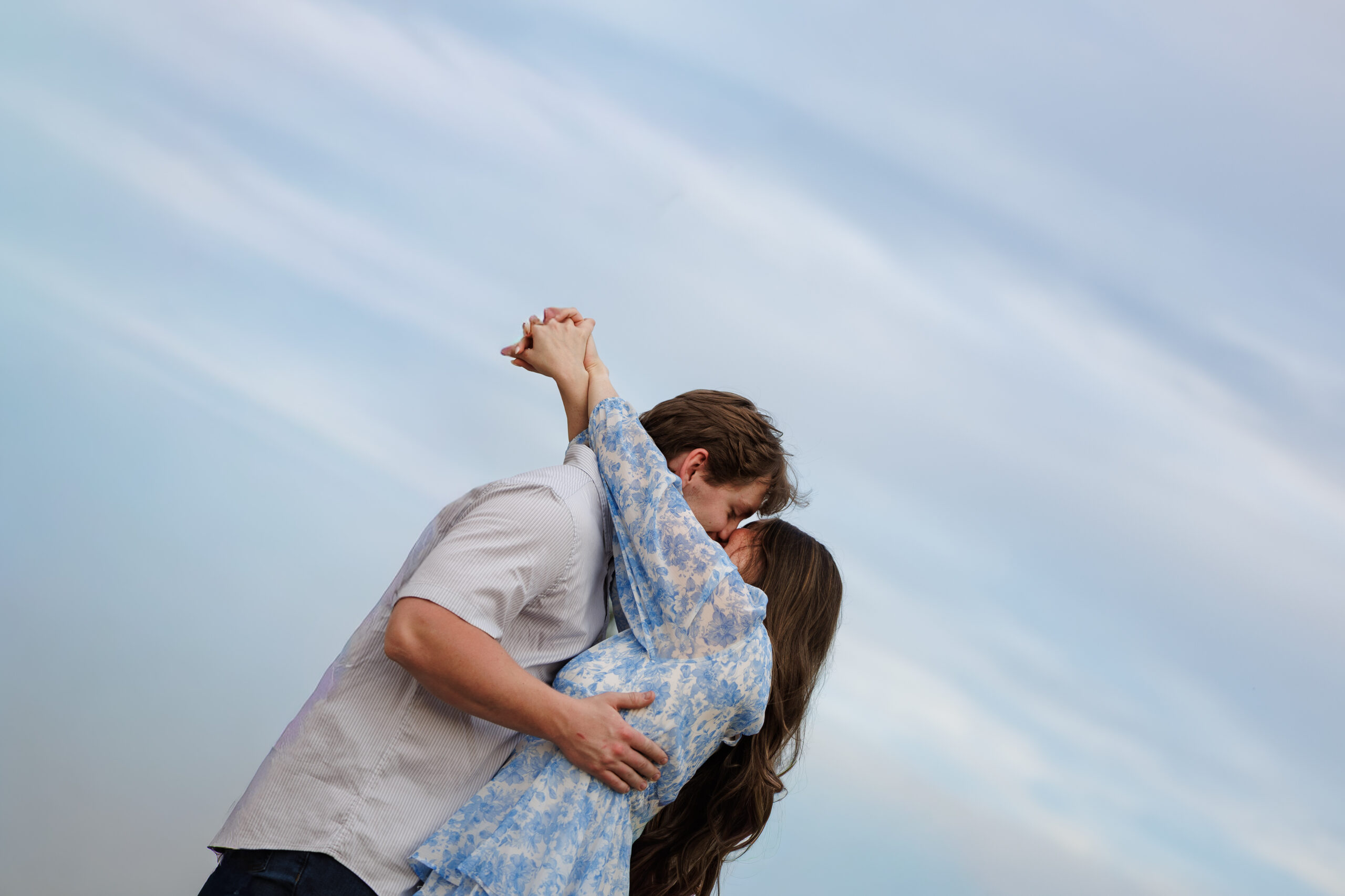 A couple laughing against a blue sky as he lifts her off her feet, from a couples session by CBK Studios.