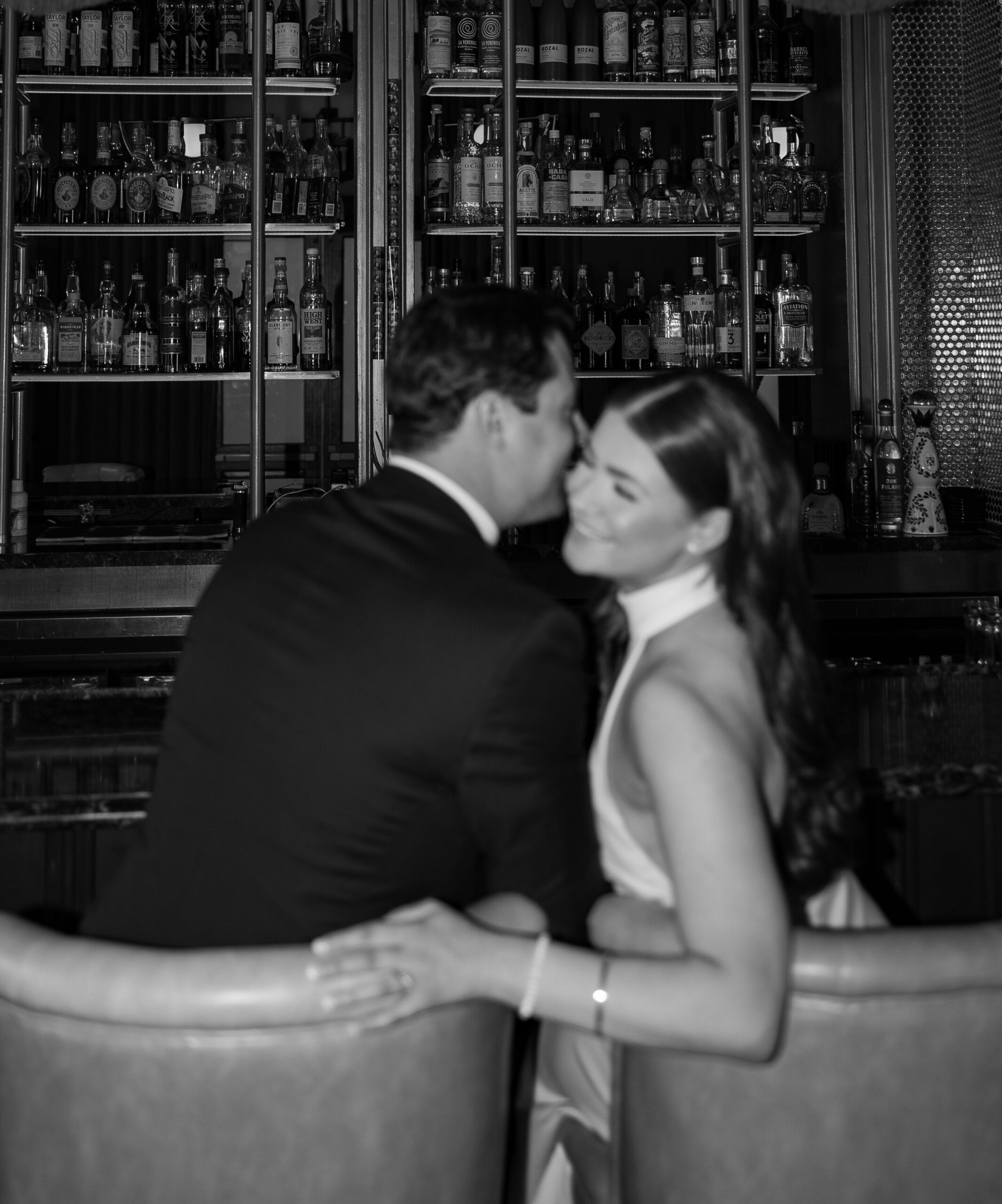 Black and white photo of a couple sharing a quiet moment at a bar, shelves of bottles behind them.