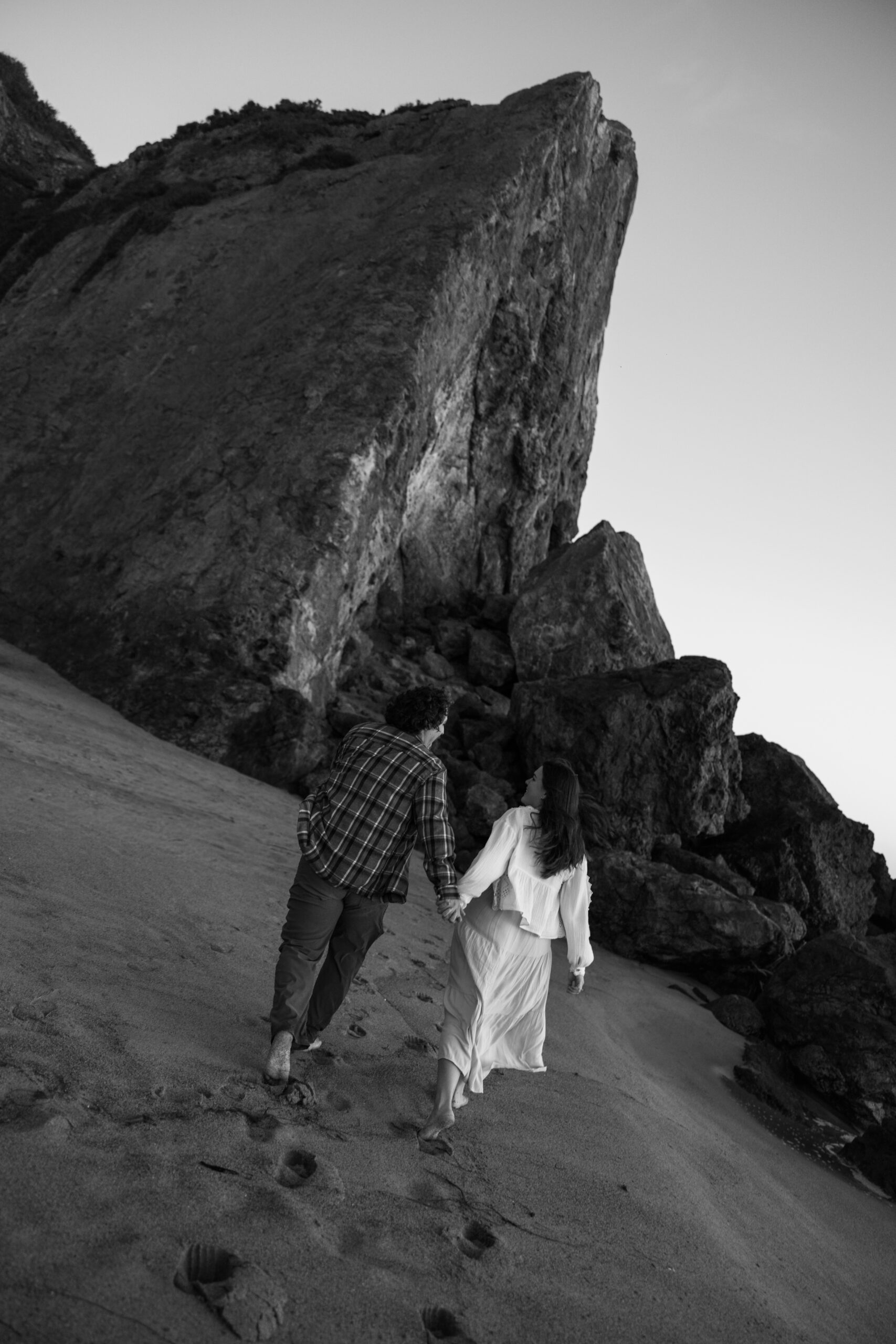 Black and white photo of a couple walking hand in hand along a sandy path beneath a rock formation.