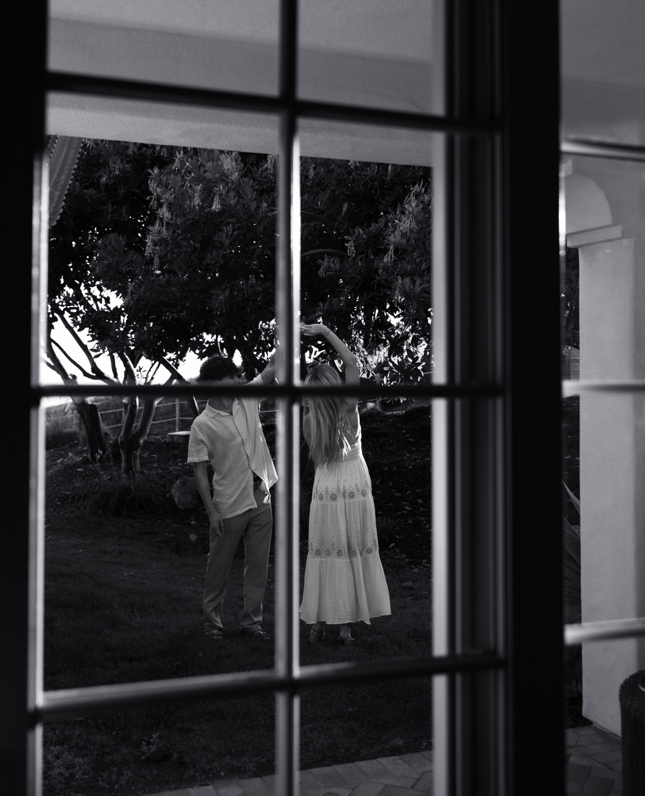 Black and white photo of a couple dancing on a lawn, seen through the panes of a window.