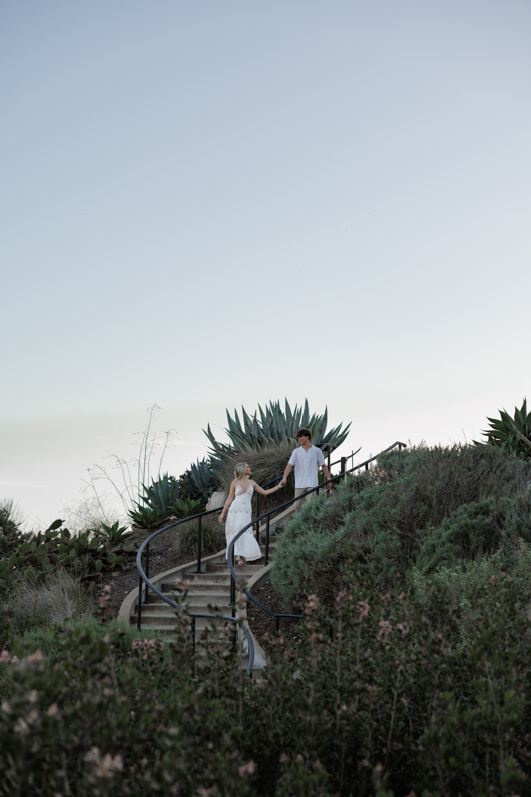 A couple walking down stone steps through coastal greenery at dusk, from a couples session.