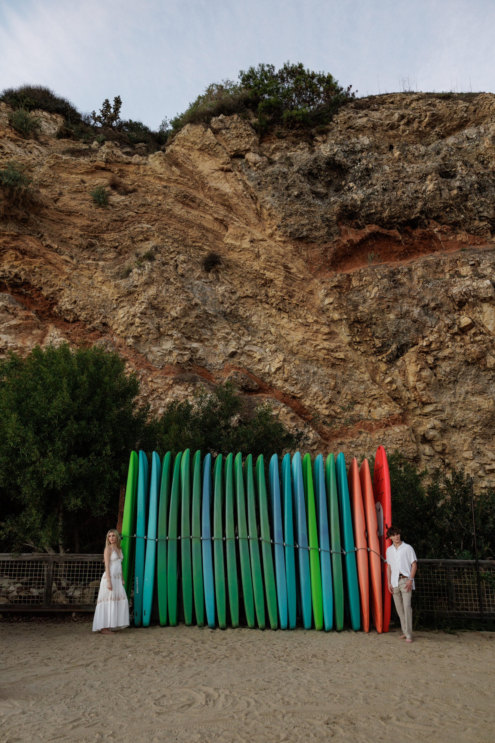 A couple standing beside a wall of colorful surfboards beneath a rocky cliff on the beach.