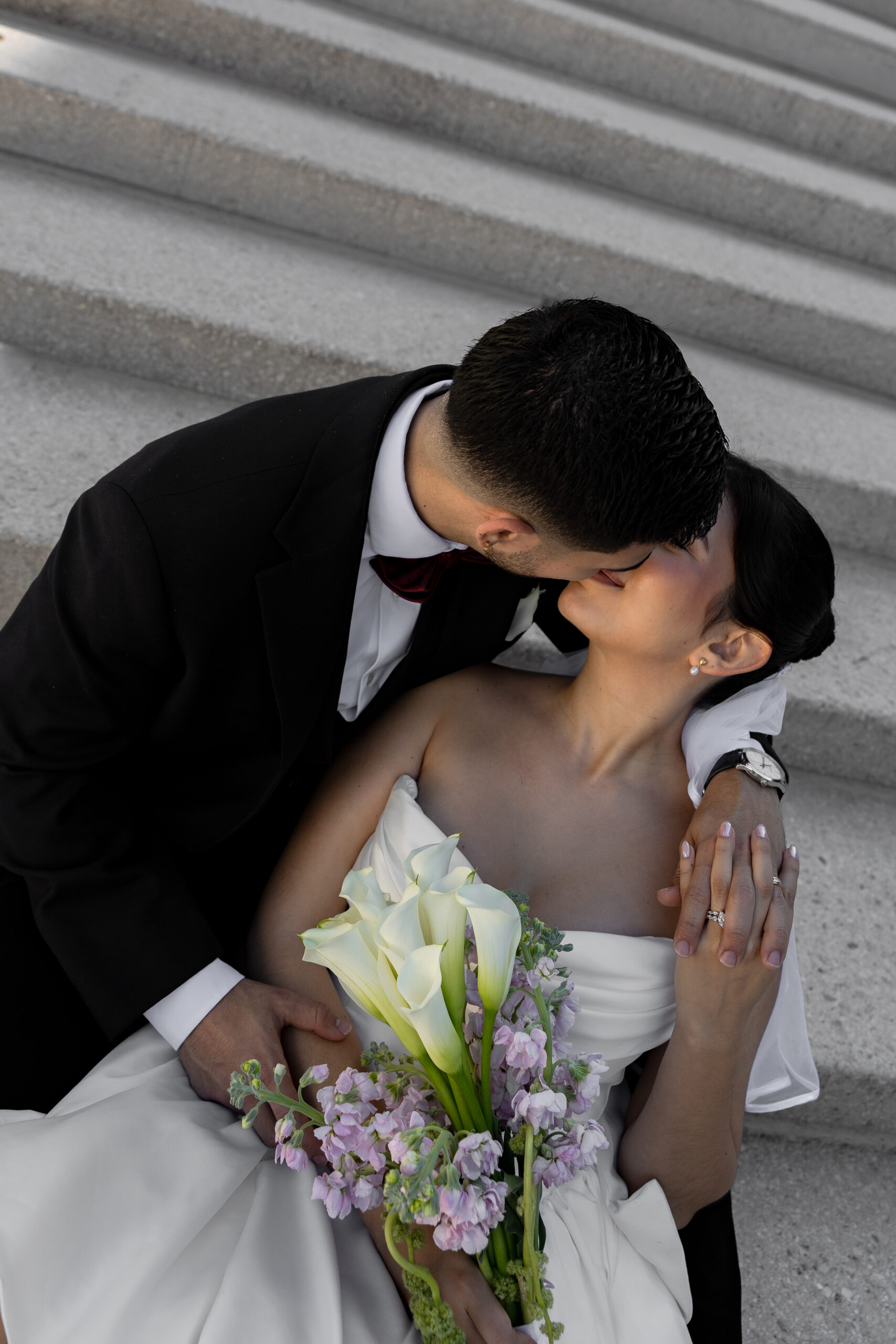 A groom kissing his bride on stone steps as she holds a bouquet of white calla lilies and lavender blooms.