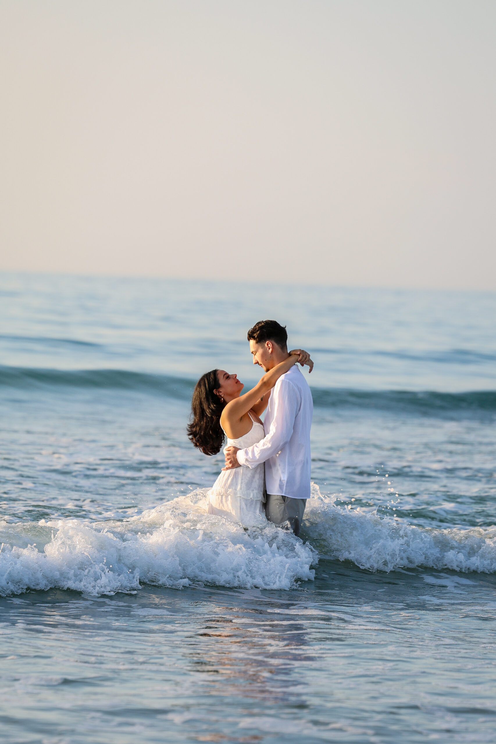 A couple in white embracing in the ocean surf with waves breaking around them.