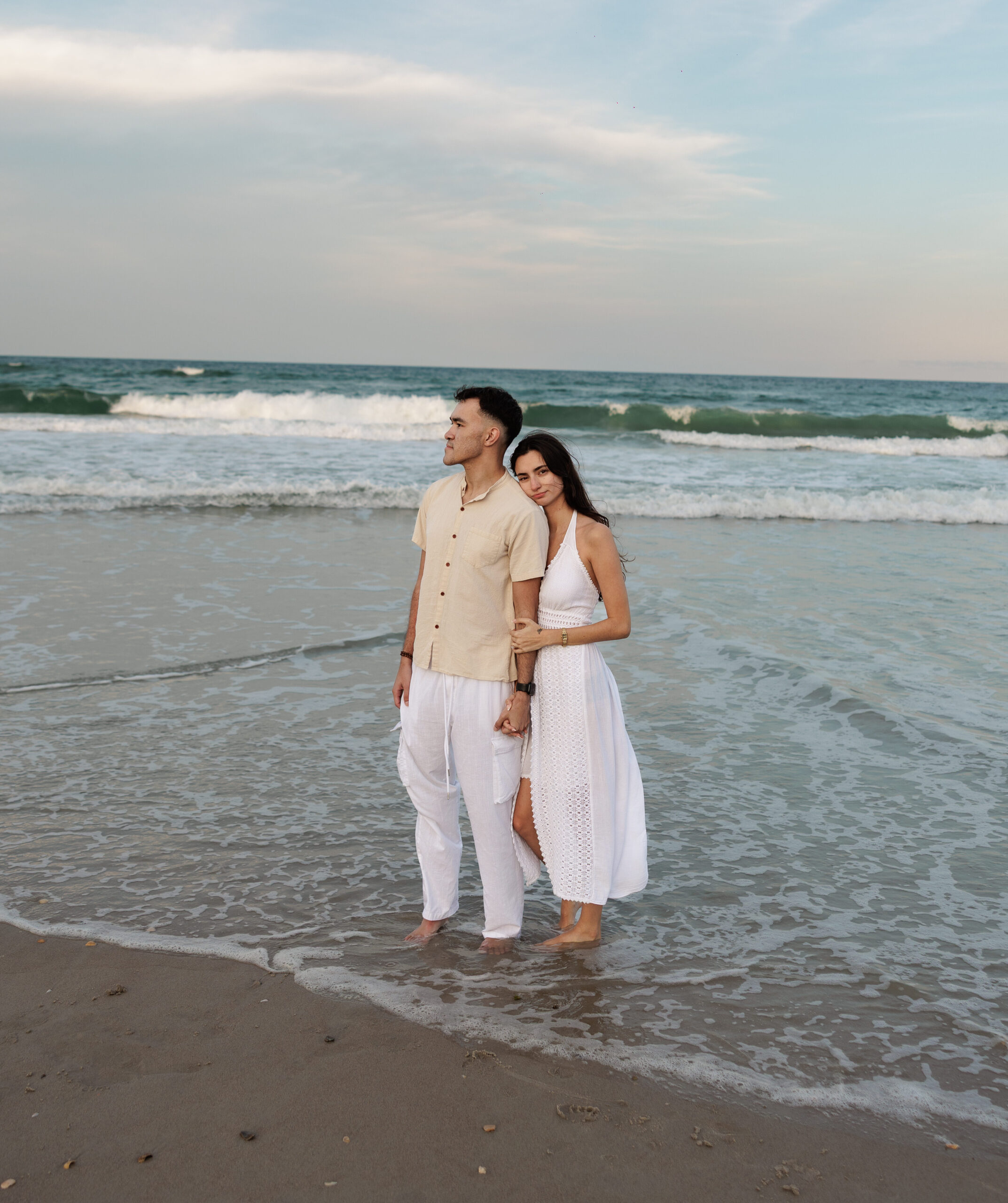 A couple in white standing at the water’s edge on the beach, waves rolling in behind them.