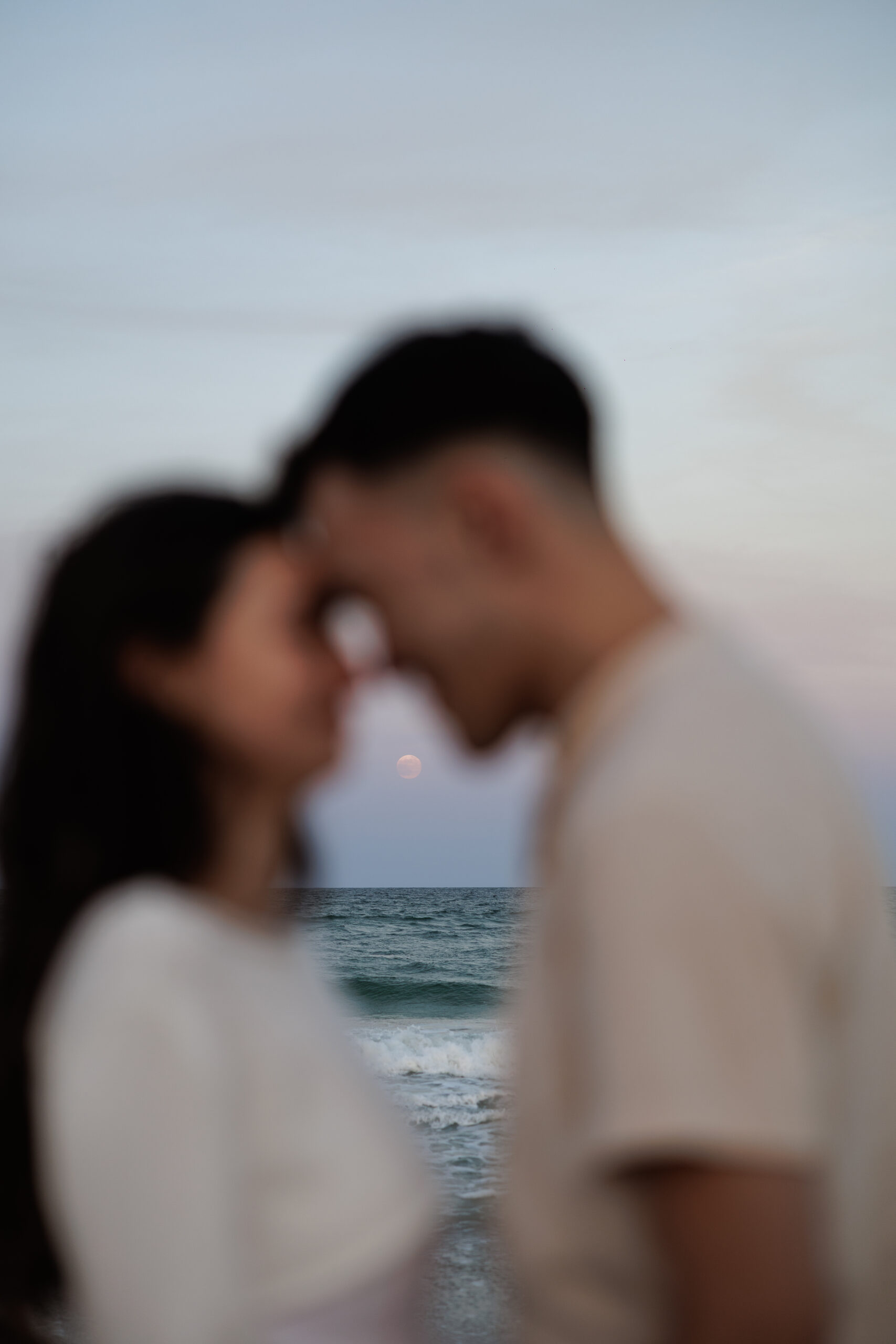 A couple forehead to forehead on the beach at dusk, softly out of focus, with the ocean behind them.