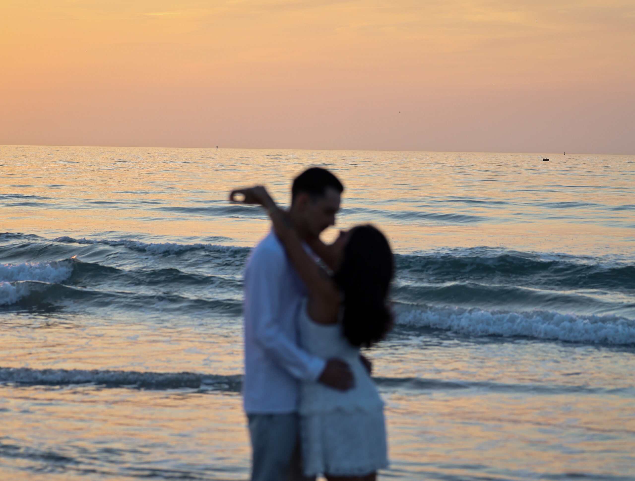 A couple embracing at the water’s edge under a pink and orange sunset sky.