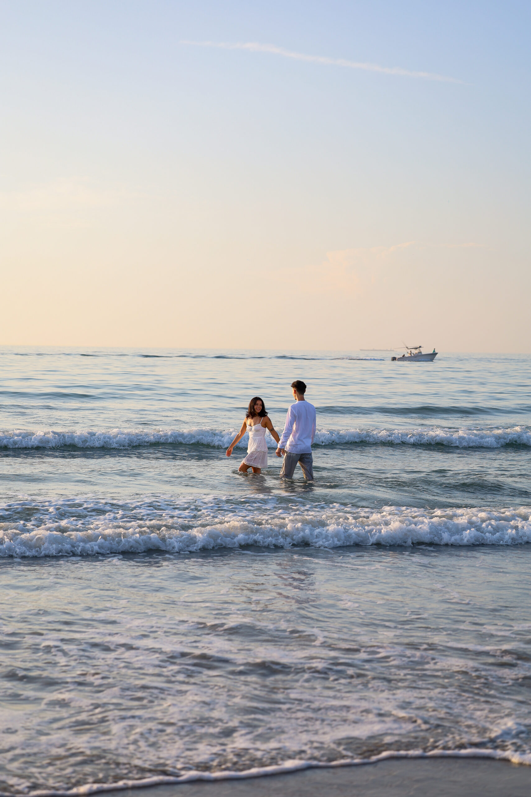 A couple holding hands as they wade into the ocean at sunset, a boat on the horizon.