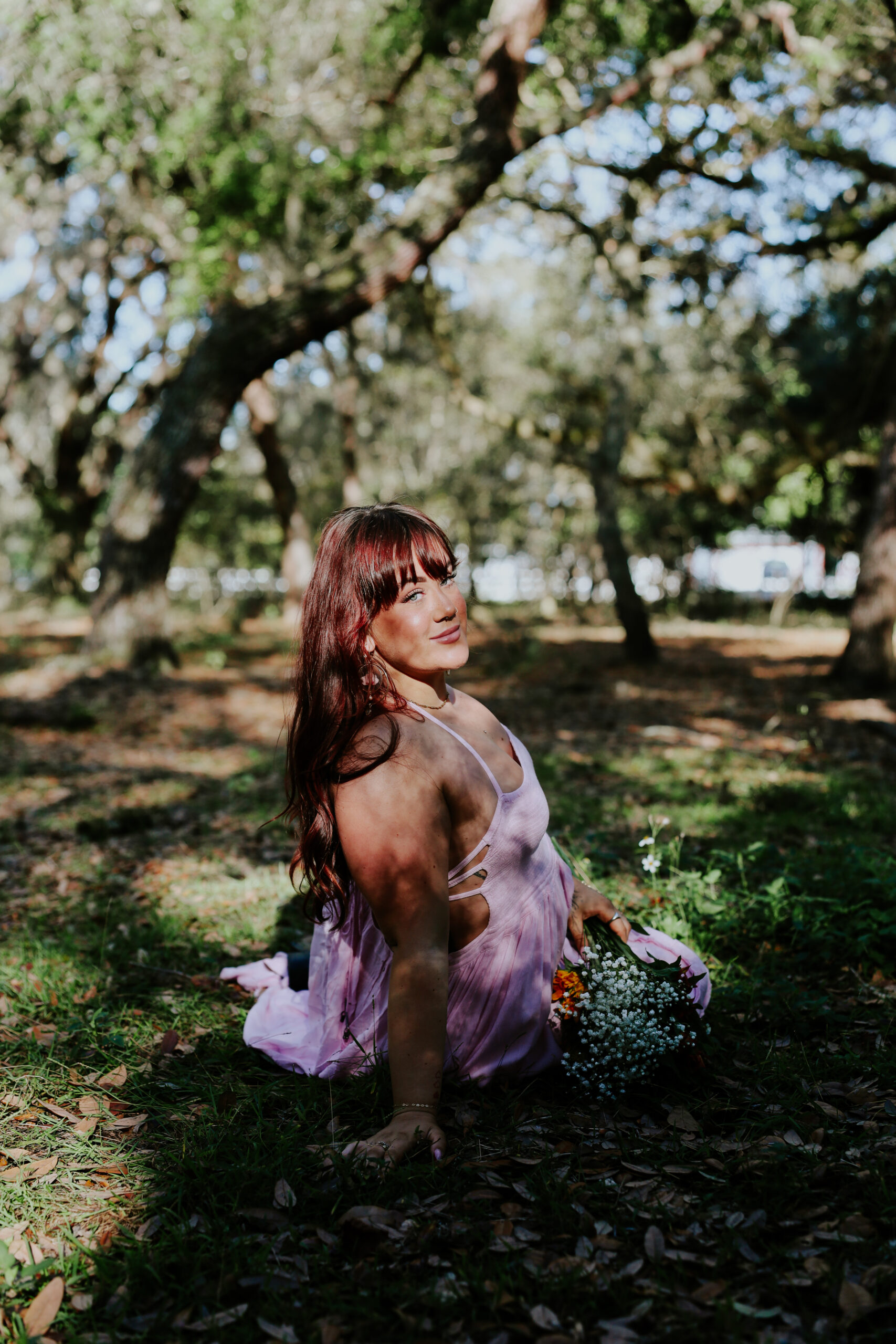 Jocelyn seated in the grass under the oaks in a lavender dress, holding a small bouquet, from her portrait shoot with CBK Studios.