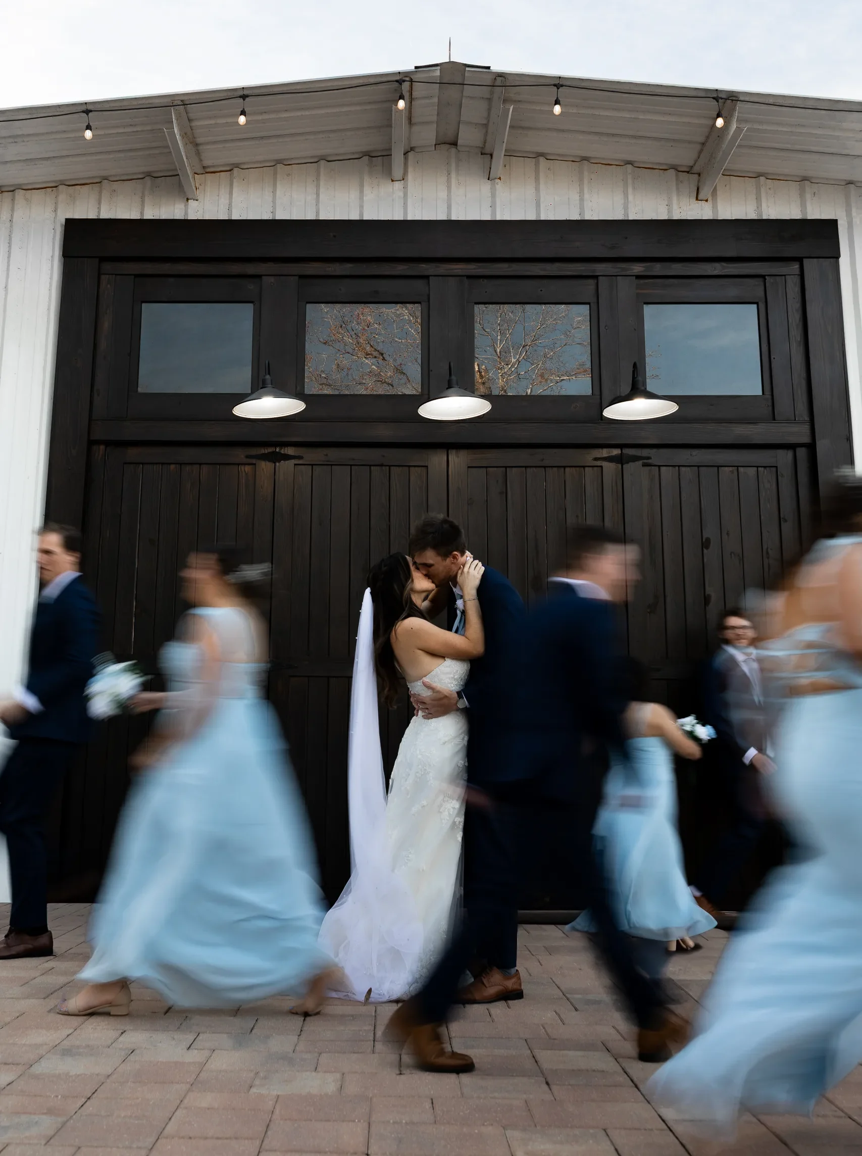 Bride and groom kiss outside the black barn doors as the wedding party passes at Cypress Creek Farmhouse, Orlando FL