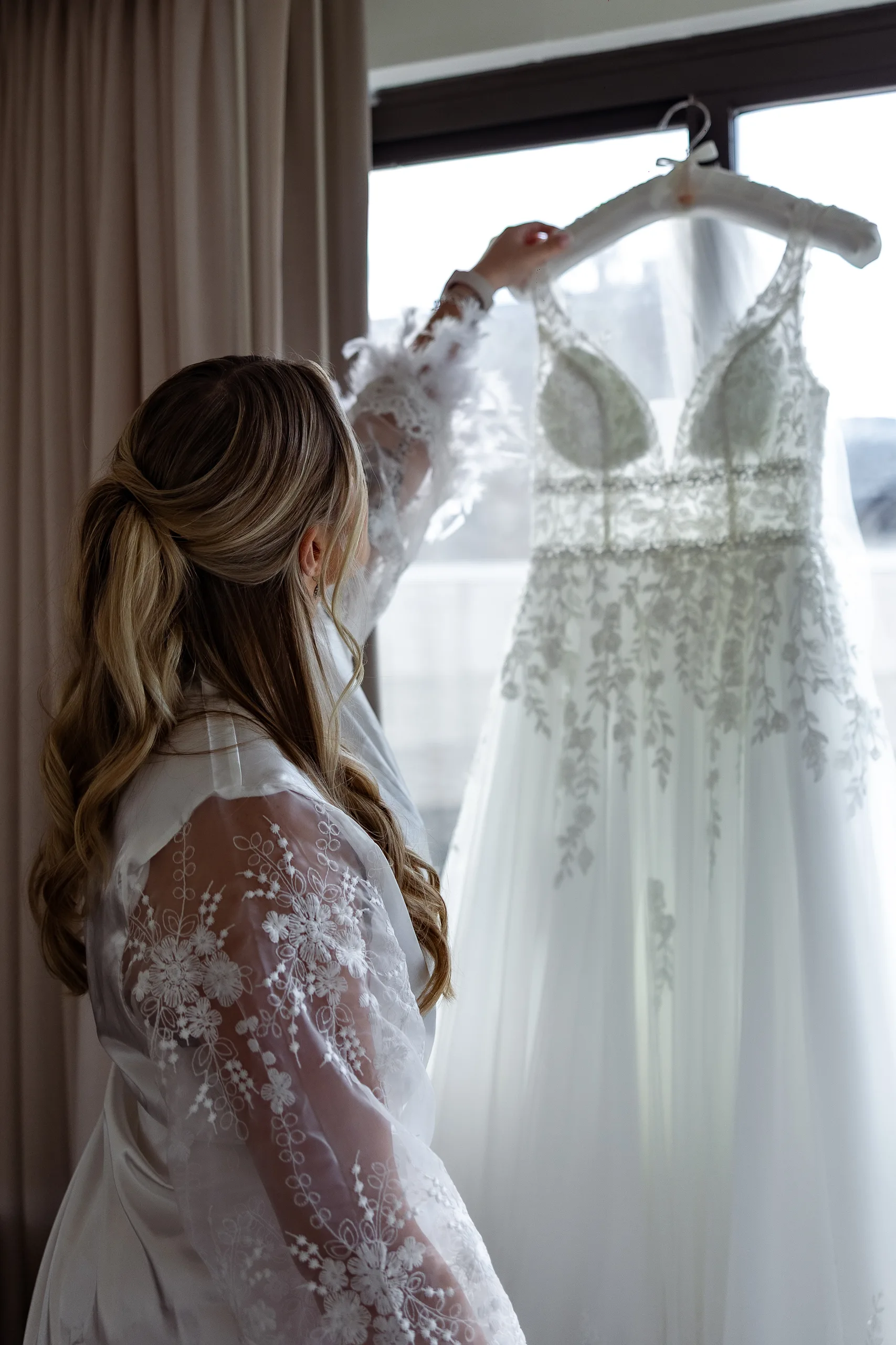 Bride in a lace robe reaching for her gown in the window light on her J House Greenwich wedding day