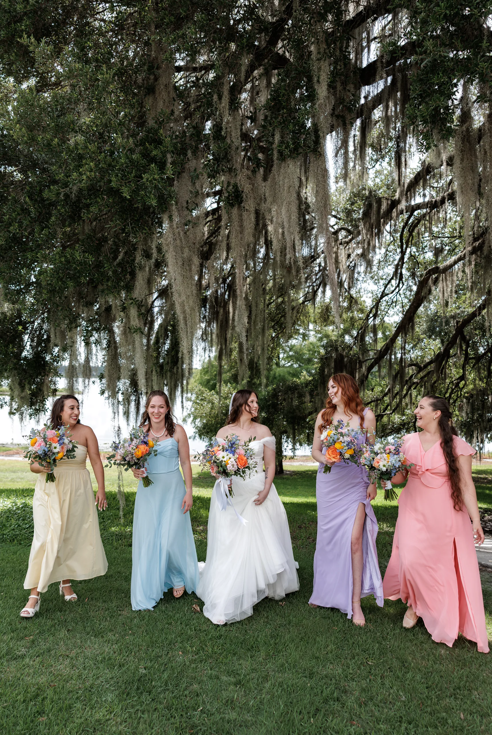 Bridesmaids in pastel dresses walking together under an oak tree at Lake Mary Events Center